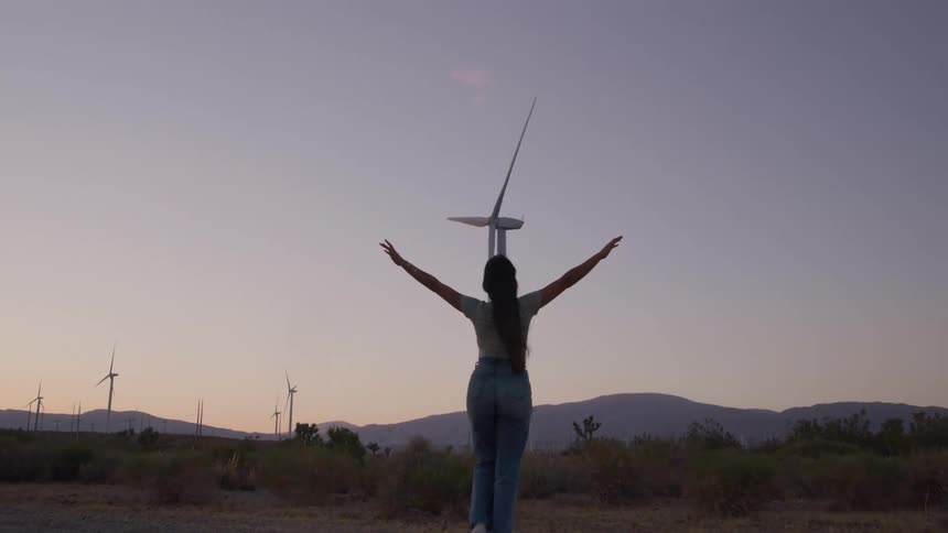 The video shot at dusk shows a woman walking away from the camera in a desolate hilly landscape dotted with wind turbinesshe stops faces a tall wind turbine and raises her arms out wide in a gesture of freedom or exhilaration against the fadin