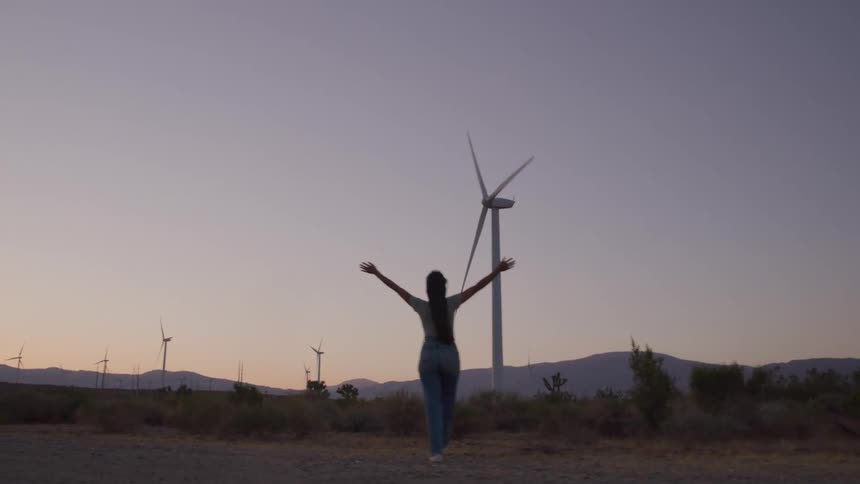 At dusk a woman walks toward a tall wind turbine with arms raised radiating carefree excitement in the open desert