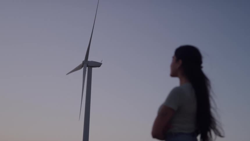 The video shows a woman seen from the side in silhouette standing with her arms crossed while looking up at a large wind turbinethe turbines blade slowly spins against the dusk sky creating a peaceful contemplative scene focused on renewab