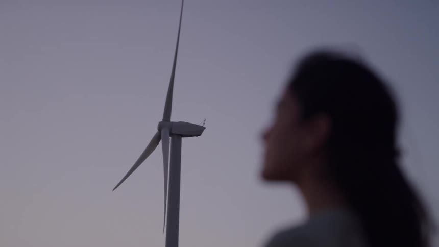 At dusk a woman in soft focus looks on as a wind turbine turns against a pale sky the scene calm and contemplative