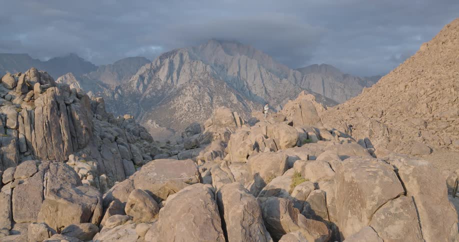 In golden early light a lone hiker stands atop pale boulders framed by dramatic granite peaks looking small