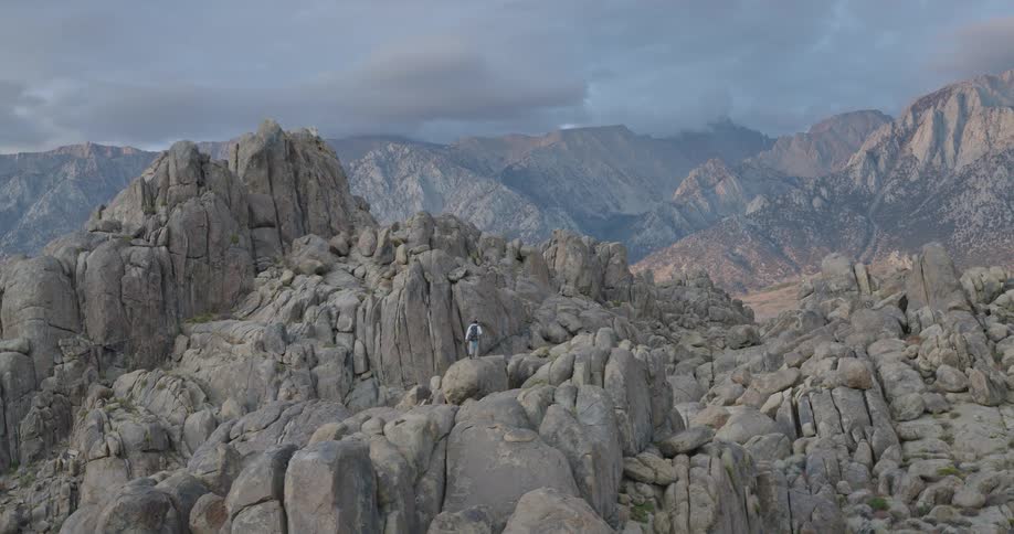 A wide view of rugged gray boulders reveals a lone hiker amid a vast mountain range under moody clouds