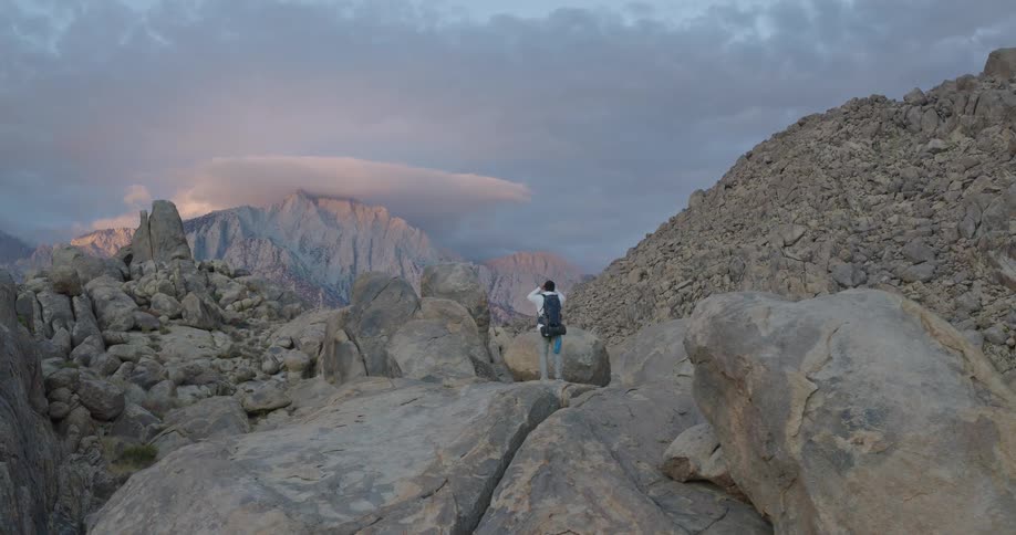The video shows a single hiker with a backpack standing amidst a vast rugged landscape of large gray boulders and rocky mountainsthe person pauses to take a photograph of the dramatic vista which includes distant towering peaks and an unusual lo