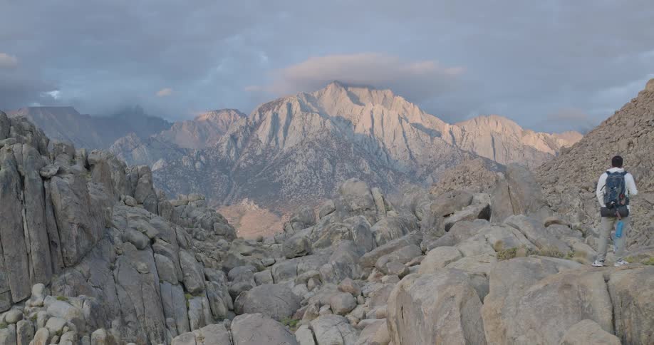 A person wearing a backpack walks along an uneven formation of large rocks toward a prominent tall mountain in the distance with the scene illuminated by lateday light under a partially clouded sky