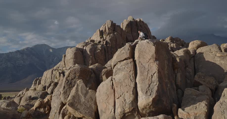A hiker with a backpack sits atop a dramatic rock formation surveying the mountainous landscape under a cloudy sky