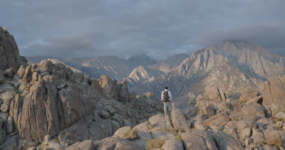 The video features a lone hiker with a backpack standing on a high point amidst a chaotic landscape of light gray cracked bouldersthe camera slowly zooms out revealing the vast scale of the rugged terrain and the imposing distant mountain