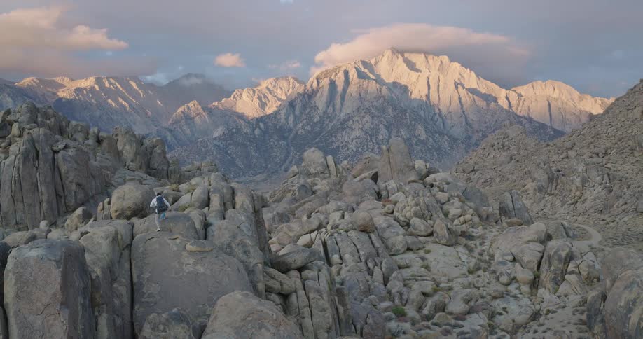 A lone hiker with a backpack traverses a vast rocky landscape with sun kissed mountains in the background