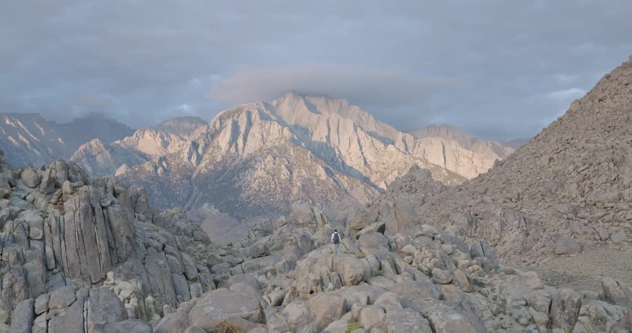 A person stands on an accumulation of rocks in the foreground facing a massive mountain range in the background under a sky with visible cloud formations