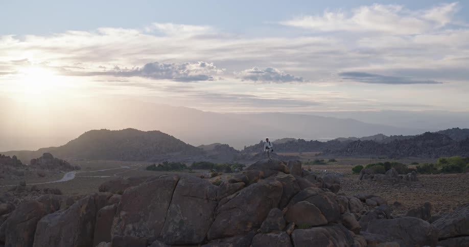 The video is a sweeping wideangle shot of a lone hiker standing on a high outcrop of large rounded bouldersthe person is admiring the breathtaking view of a vast desert landscape that stretches out below and is framed by distant hazy mounta