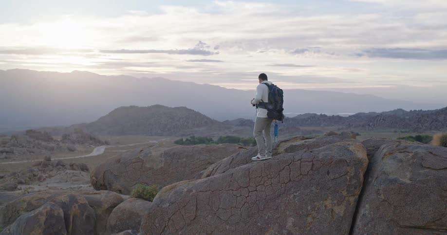 The video shows a male hiker wearing a backpack standing on a high point of large cracked rock formationshe is holding and looking through binoculars at the vast hazy landscape stretching out before him suggesting he is taking a break to obs