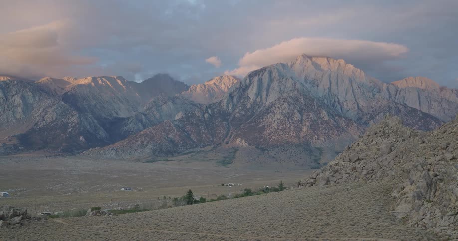 Soft evening light bathes a rugged granite mountain range as lenticular clouds hover above a wide sagebrush valley with a few scattered homes