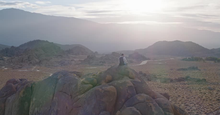A lone hiker sits atop a large rounded rock formation in a vast arid landscape gazing out at the distant mountains shrouded in a hazy or misty atmosphere the camera slowly zooms in emphasizing the scale of the environment and the contemplative nature