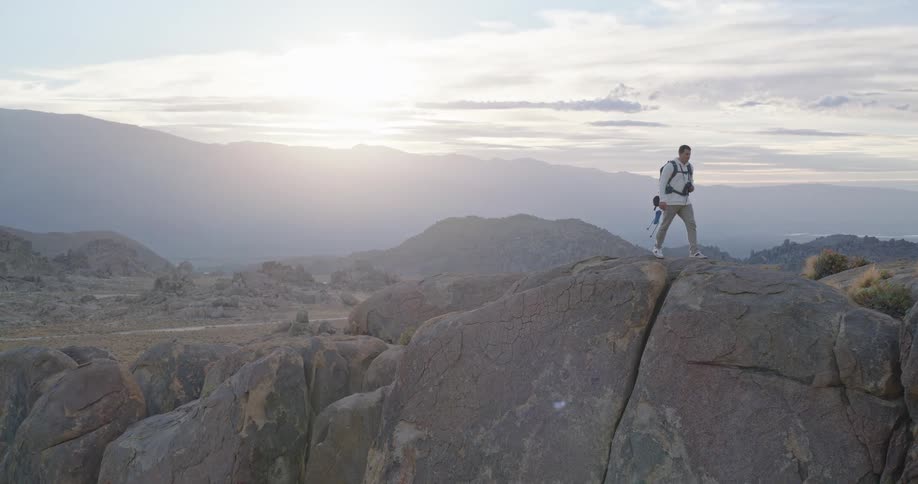 A male hiker wearing a white shirt khaki pants and a backpack stands on the edge of a large cracked boulder formation looking out over a vast hazy mountain landscape where the sun is setting or rising in the background