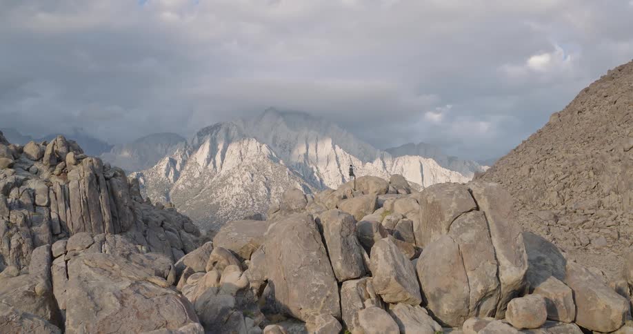 A wide view of rugged gray boulders with a lone hiker amid a vast mountain range under moody clouds