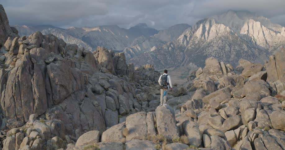 A solo hiker walks then stands among sprawling tan boulders dwarfed by a dramatic wall of granite peaks under brooding clouds appearing focused and small against the vast landscape