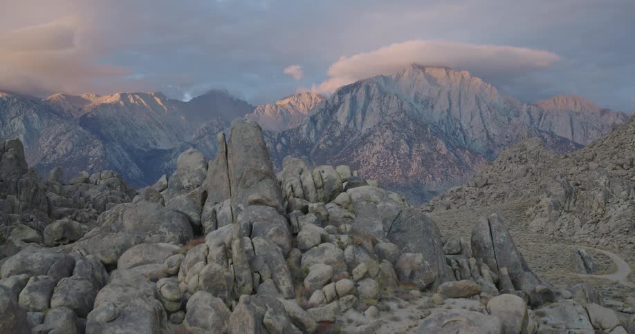 An aerial view shows a lone backpacker moving across a rugged field of large irregularly shaped boulders and rock formations the landscape is dominated by the dramatic backdrop of tall jagged mountains under a partly cloudy sky