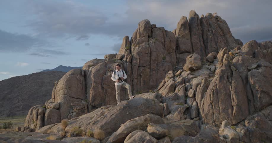 A lone man stands atop sunlit granite boulders scanning a rugged mountain range through binoculars beneath brooding clouds appearing focused and calm