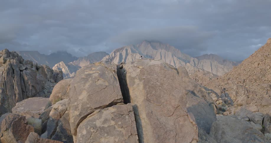 The video is an extreme long shot of a man hiking alone among the large smooth gray boulders and rock formations of a mountain or desert landscapethe camera slowly pulls out and up making the hiker look small as the vast desolate and rugge