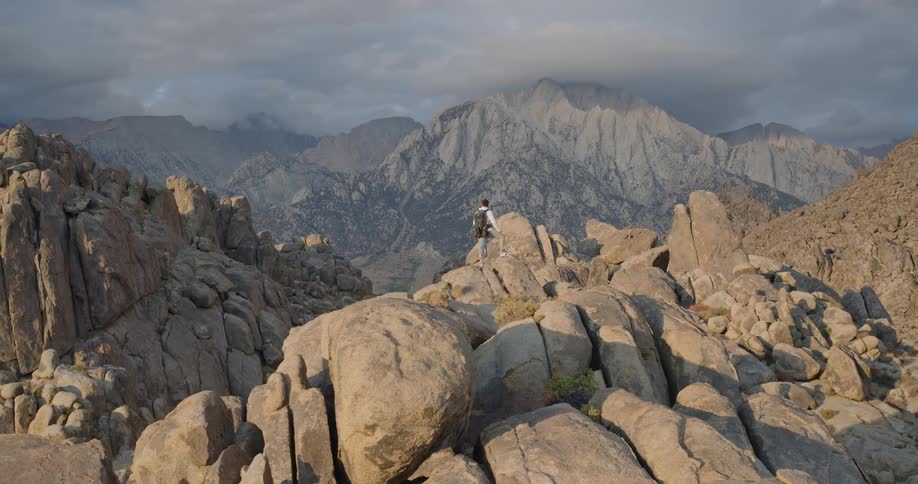 The video is an extreme long shot showing a hiker standing on a large prominent rock formation in a desolate mountain landscapethe camera slowly pans to the right emphasizing the vast scale of the rugged rocky terrain and the imposing hazy mo