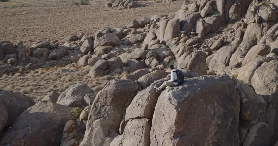 A man with a backpack sits on a large prominent rock in a vast area of scattered boulders and dry ground as the camera ascends to reveal a distant towering mountain range beneath a cloudy sky