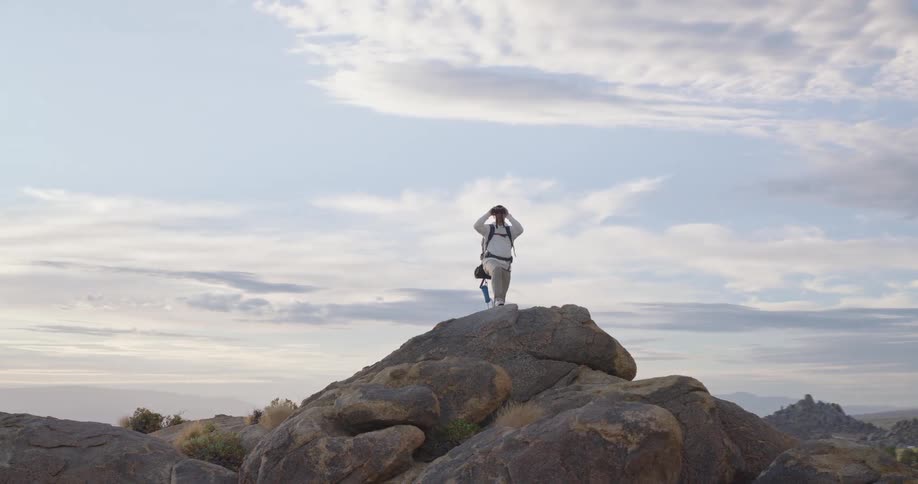 A hiker with a backpack stands on a rocky summit looking through binoculars at the vast mountain landscape