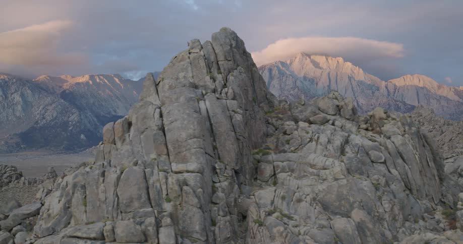 A dramatic sweeping camera movement reveals a landscape dominated by massive rugged grey boulders in the foreground and high jagged mountains in the background at sunset a cloud casts a soft ethereal glow on the highest peaks hinting at the vastness