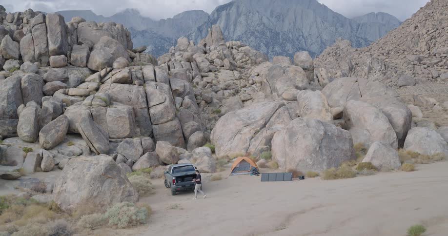 A lone camper stands beside a pickup an orange tent and a row of portable solar panels tucked among towering rounded boulders