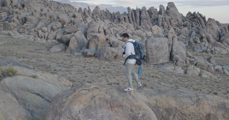 The video is an extreme long shot that slowly pans out showing a hiker standing on a high point amidst rugged rock formationsthe hiker is dwarfed by the massive scale of the desert mountain landscape which is a wash of gray and brown tone