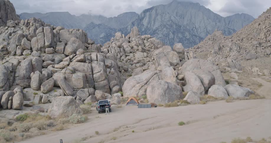 A young man carrying a backpack and holding a camera walks toward the mountain range after packing up his truck ready to explore the rugged terrain ahead