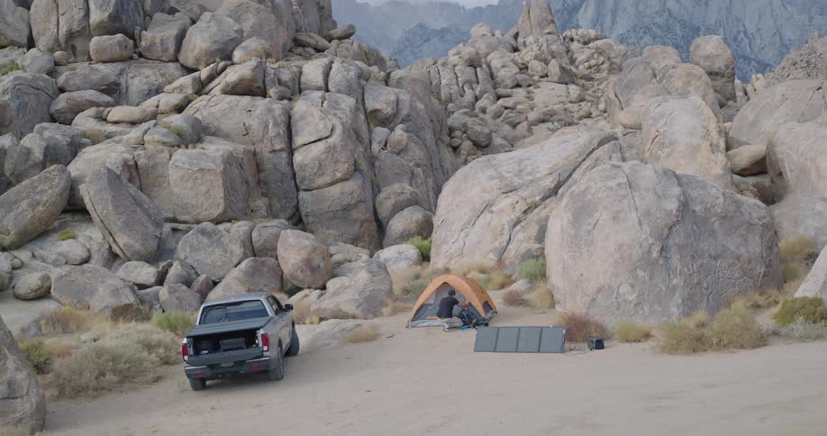 A pickup truck is parked near an orange tent and an unfolded array of solar panels and a person walks toward the tent and then bends down to adjust equipment on the ground all situated on a patch of dirt surrounded by massive rock formations