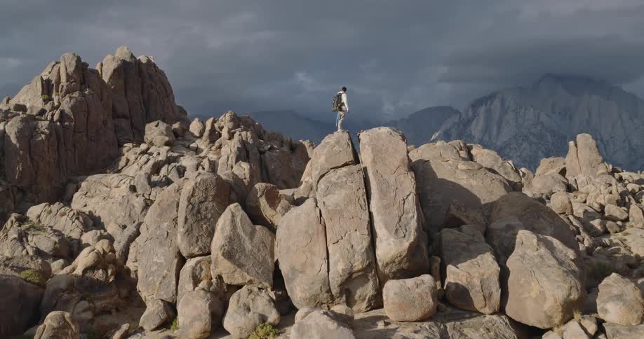 A person stands briefly atop the highest point of a cluster of boulders under a cloud filled sky