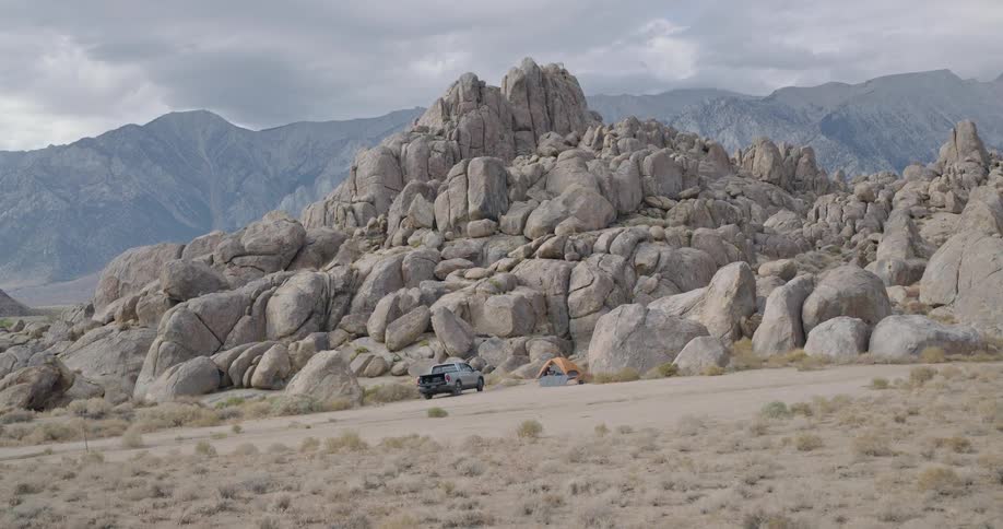 The video shows a timelapse of a rocky mountainous landscape likely the alabama hills in california under a dramatic cloudy sky a person sets up a campsite in the foreground positioning a pickup truck a tent and a portable solar panel system near