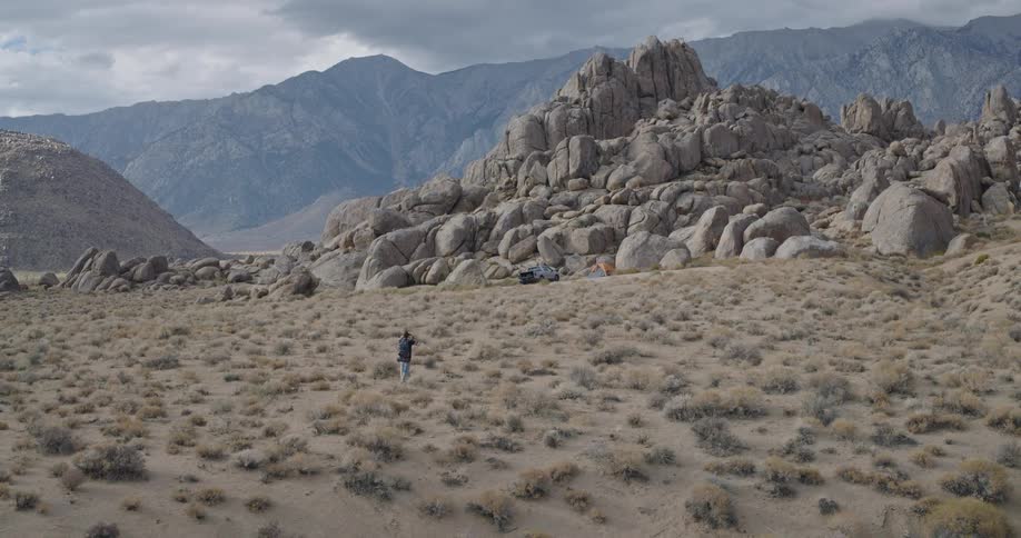 Person walking in arid terrain with mountains in the background and a campsite near the rocks