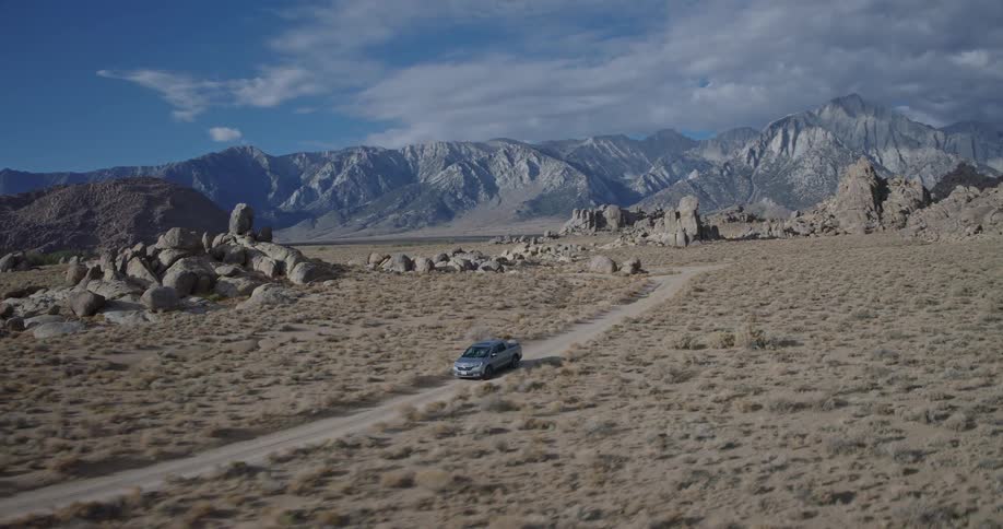 The video is an establishing shot of a vast desert landscape with a silver pickup truck driving along a dirt road in the foregroundthe background is dominated by a majestic hazy mountain range under a partially cloudy blue sky with large