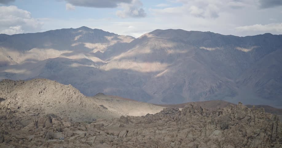 Sunlit mountains with rocky terrain in the foreground and partly cloudy sky