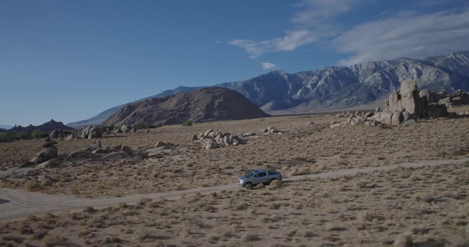 An aerial shot tracks a silver pickup truck driving on a dirt road through a vast rocky desert with a mountain range in the background