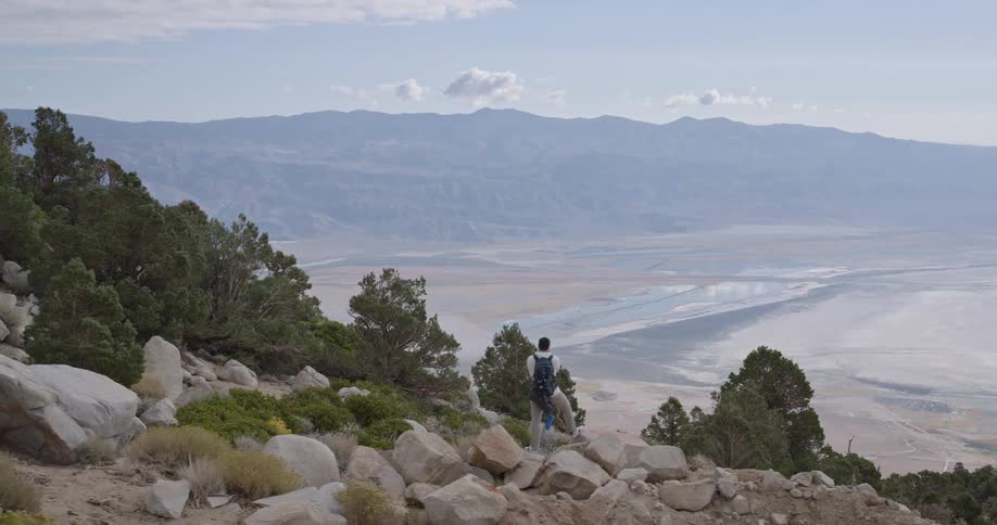 A person with a backpack stands on a rocky vegetated slope looking out over an extremely wide flat valley floor that recedes into multiple layers of distant mountain ranges under a light sky