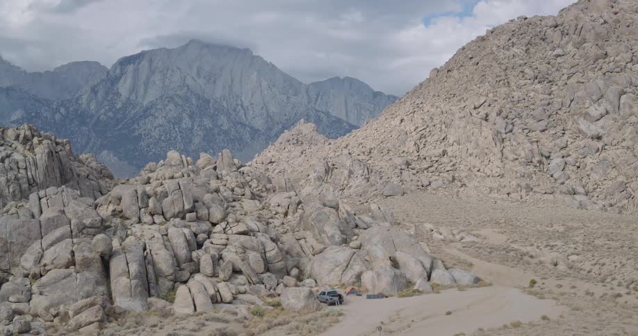 The video is an extreme long shot of a secluded campsite in a vast rocky mountain and desert landscapea dark suv or truck is parked with a tent pitched nearby completely dwarfed by the massive scale of the rugged graybrown terrain