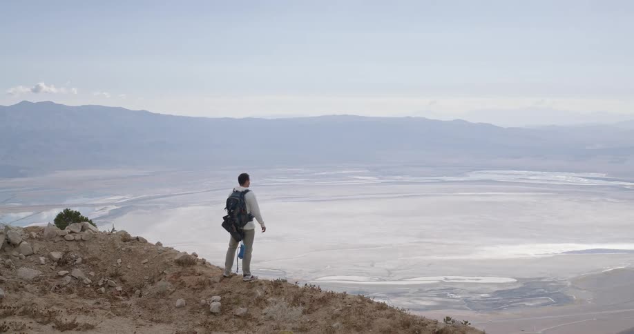 A hiker with a prosthetic leg and a backpack stands on a mountainside looking out at a vast arid valley below