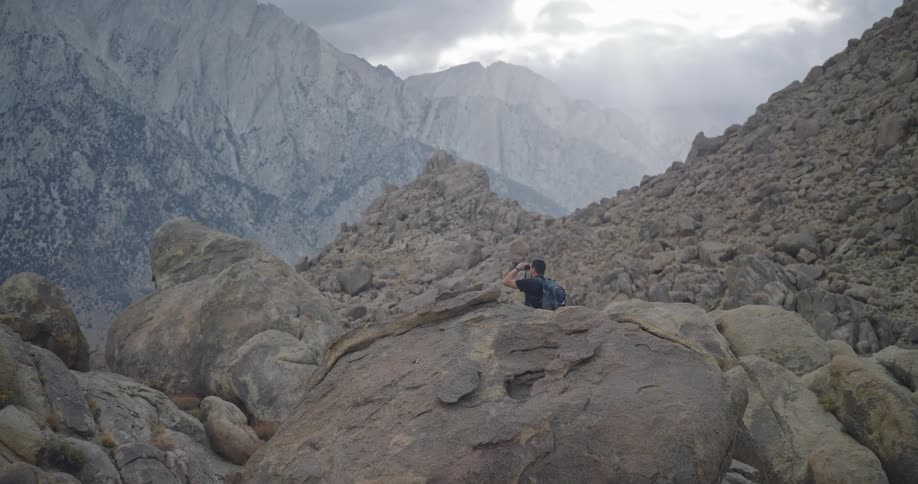 Man standing up looking at a mountain range through binoculars outdoors during day time