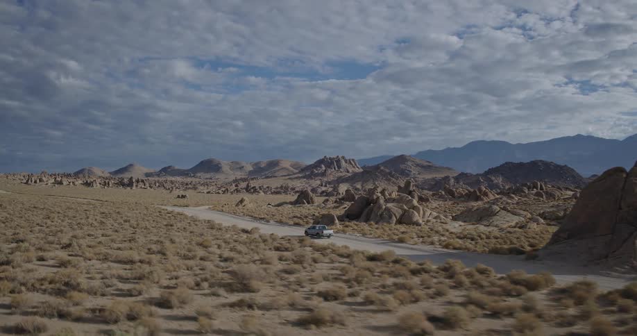 A silver pickup truck drives on a dirt road through a vast rocky desert landscape under a dramatic cloudy sky