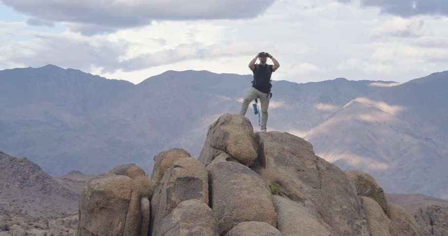 A man with a backpack stands atop a rocky outcrop using binoculars to scan the vast mountain landscape under a partly cloudy sky