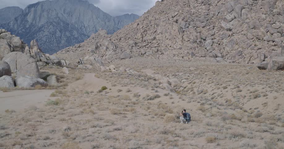 A man with a backpack crouches in a scrubby desert basin beneath a rocky mountainside peering through binoculars with a focused curious expression
