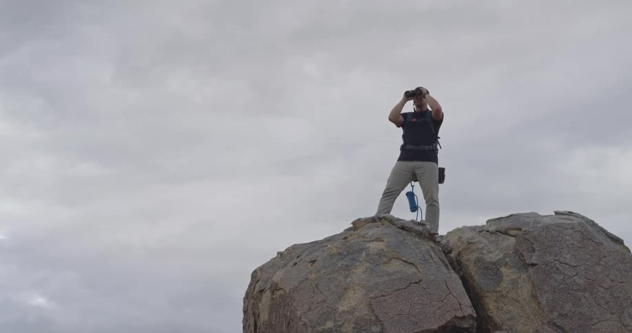 Man with backpack using binoculars atop rock formation in mountainous area under cloudy sky