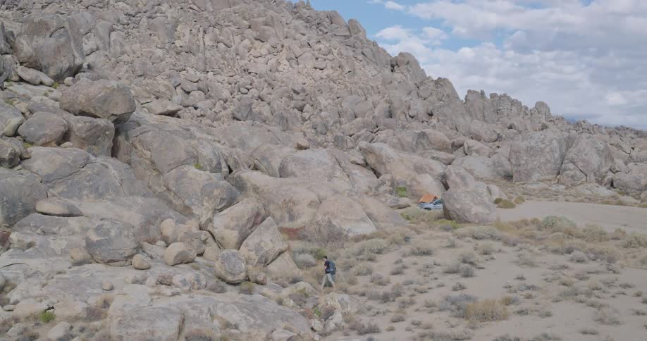 A lone hiker with a blue backpack walks across a scrubby desert valley beneath towering granite ridges