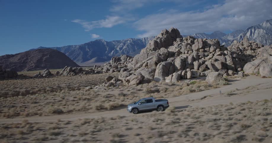 A silver pickup cruises along a sandy desert track past a cluster of smooth rock formations under a bright blue sky with scattered clouds