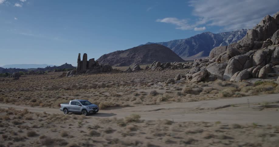 A gray pickup truck drives slowly across a dirt road through a wide sparsely vegetated desert valley passing large clusters of boulders with imposing mountain ranges visible in the distance under a partly cloudy sky