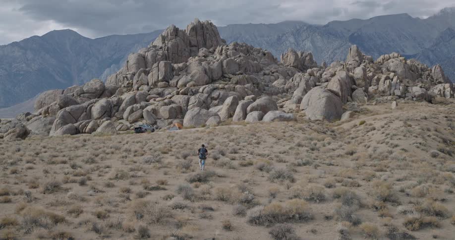 A lone hiker strides across a wide scrubby desert toward towering rounded granite outcrops with blue gray mountains under a cloudy sky
