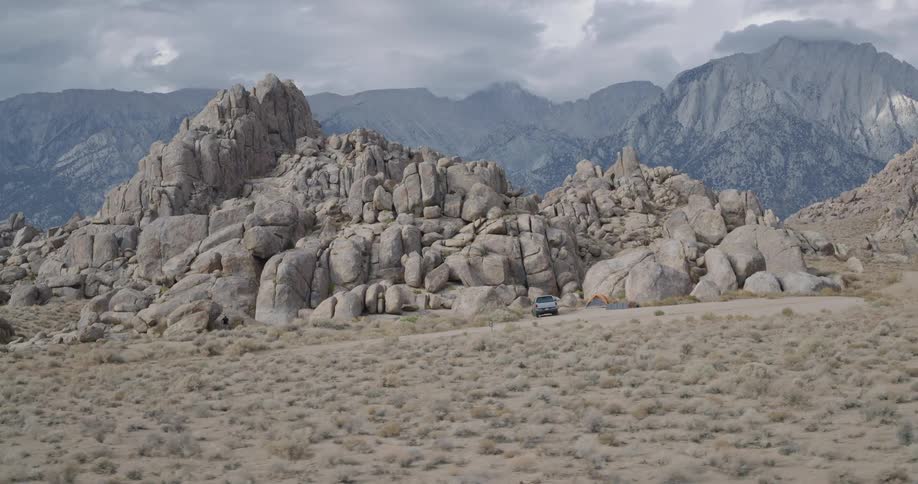 A gray pickup truck drives to a stop near an orange tent pitched beside a cluster of massive rounded rock formations all situated in a wide arid valley with tall dark mountains visible on the distant horizon under an overcast sky