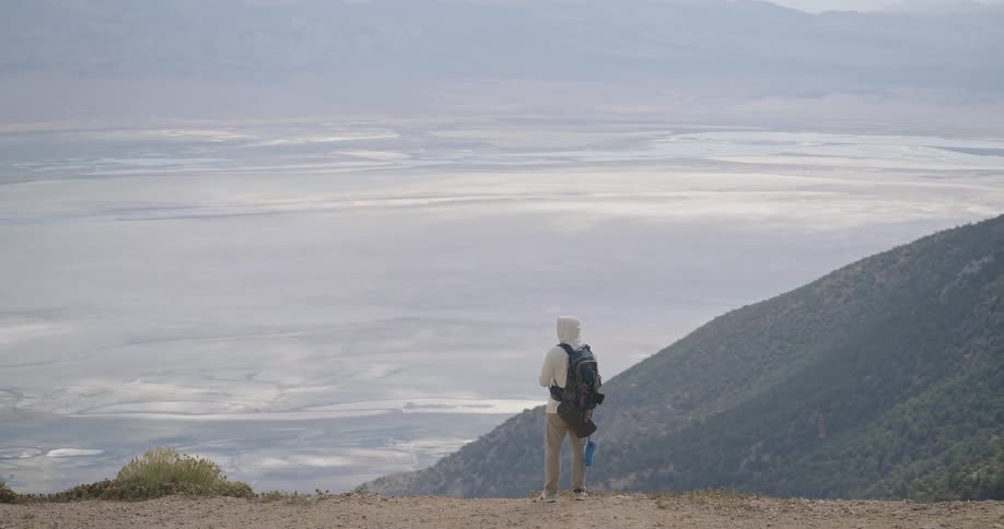 A person with a backpack stands on a wide dirt ledge at the top of a steep vegetated hill using a device to photograph a massive pale valley floor that stretches to distant hazy mountains